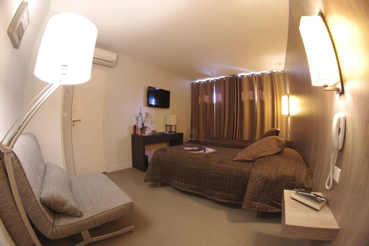 Ein Hotelzimmer mit einem Bett, einem Nachttisch mit Lampe, einem Sessel, einem Schreibtisch mit Fernseher und einem Telefon.