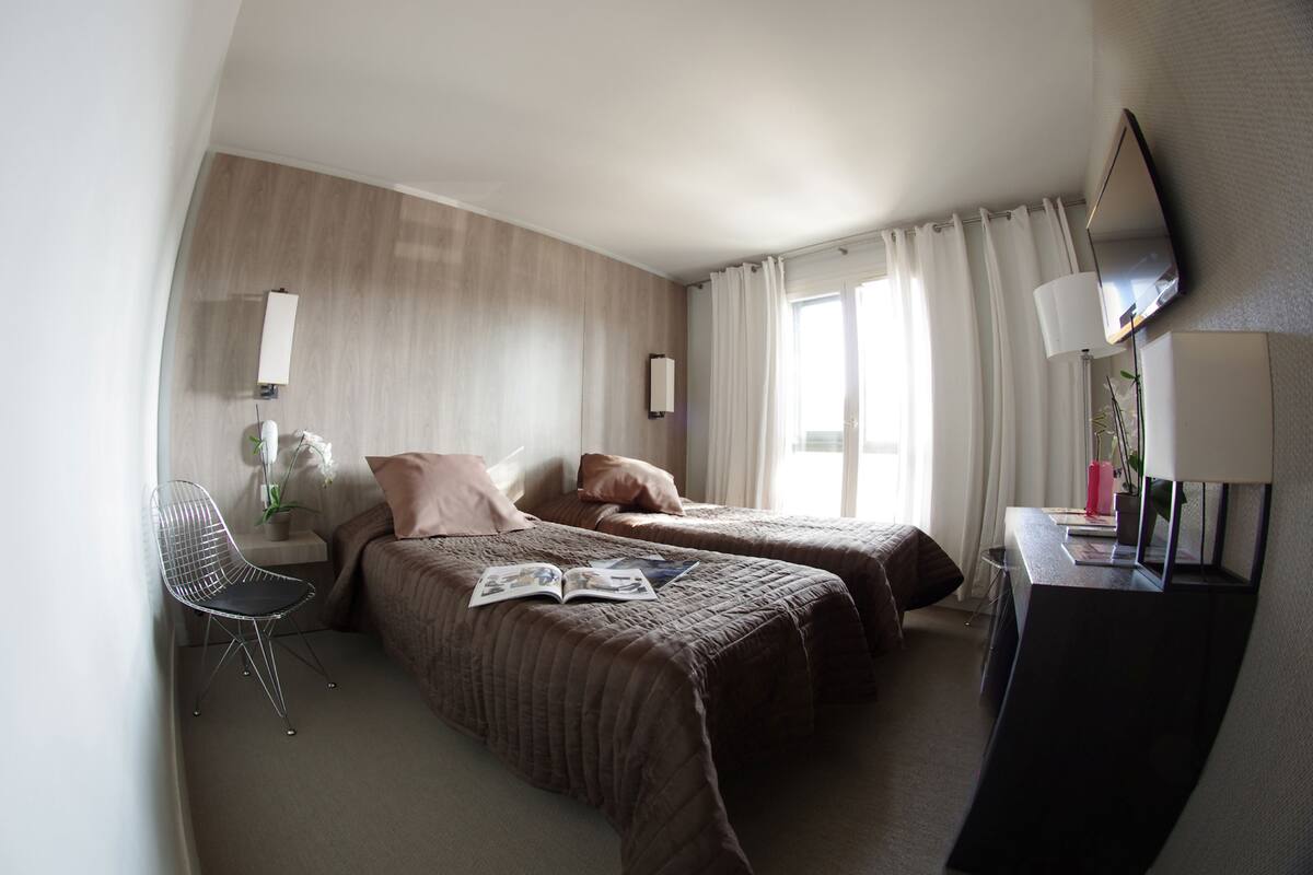 Ein Hotelzimmer mit zwei Betten, einem Schreibtisch, einem Stuhl, einem Fernseher und einem Fenster mit Vorhängen.