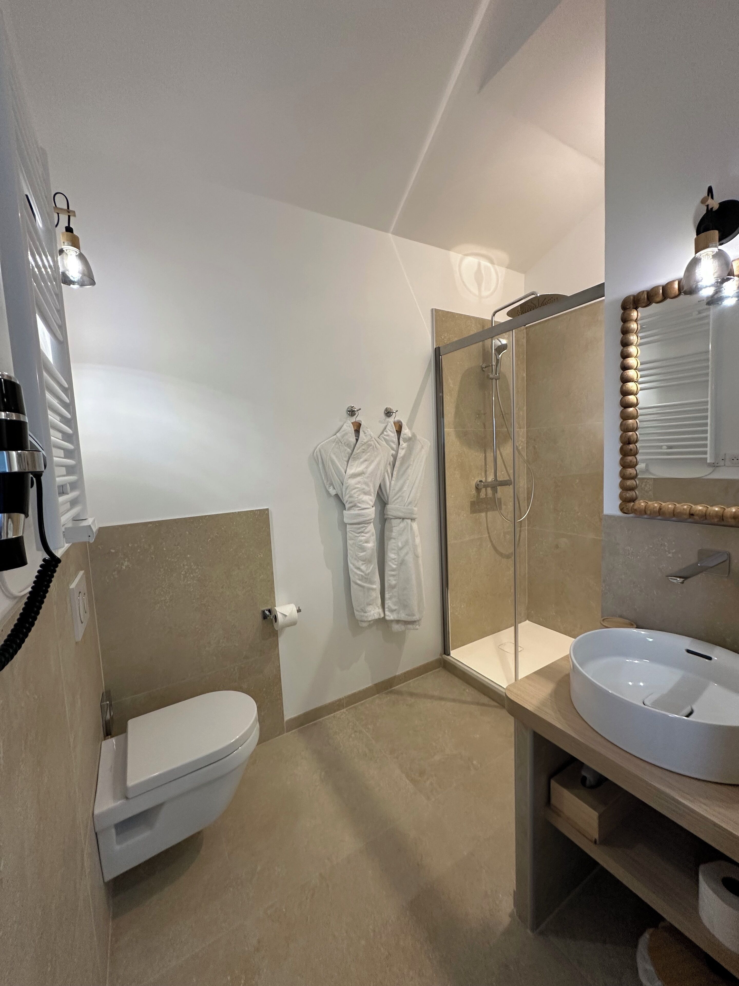 A modern bathroom with a toilet, a glass-enclosed shower, a white sink, and two white robes hanging on the wall.