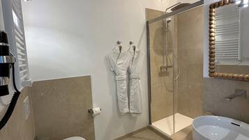 A modern bathroom with a toilet, a glass-enclosed shower, a white sink, and two white robes hanging on the wall.