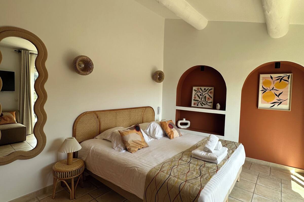 A bedroom with a bed, a bedside table, a lamp, a mirror, and a wall with decorative items.