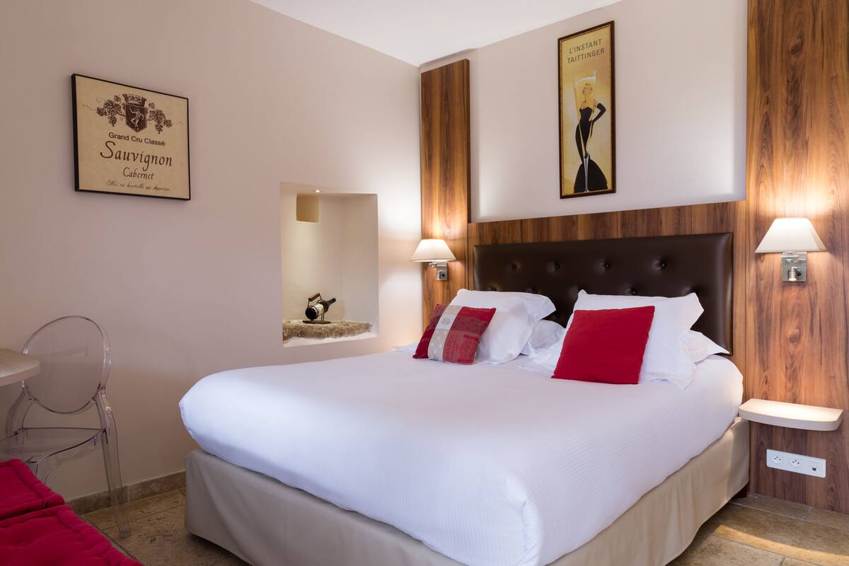 A hotel room with a large bed, a wooden headboard, two wall-mounted lamps, a framed certificate on the wall, and a small shelf with a camera.