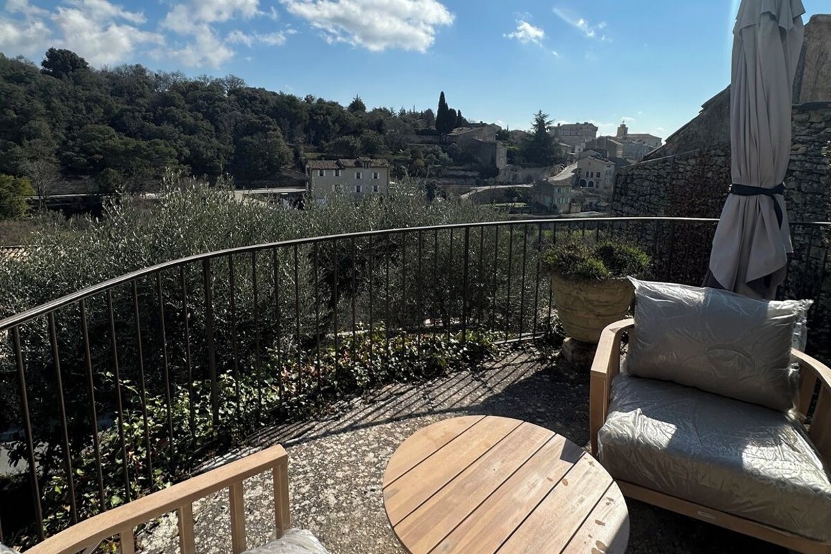 A balcony with a wooden table and a cushioned chair, overlooking a hillside with buildings and trees.