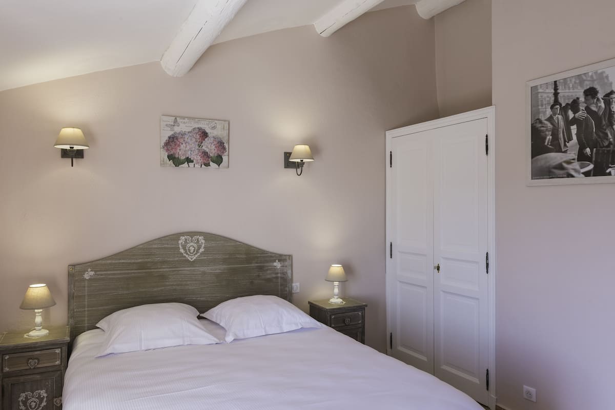 A bedroom with a wooden headboard, two bedside lamps, a nightstand with a drawer, a white door, and a framed picture on the wall.
