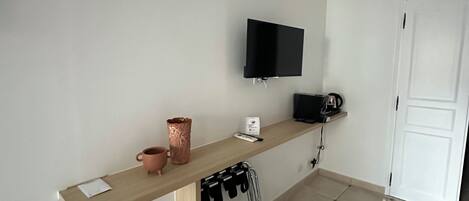 A small, clean room with a flat-screen TV mounted on the wall, a wooden shelf with a coffee maker, and a trash bin.