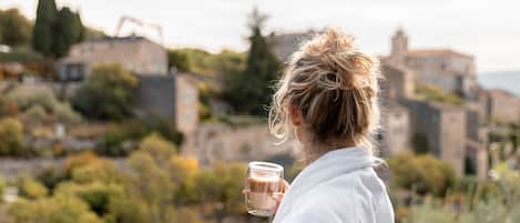 Una persona vestida con una bata blanca sostiene un vaso de bebida, observando una vista panorámica de un pueblo con edificios y vegetación.