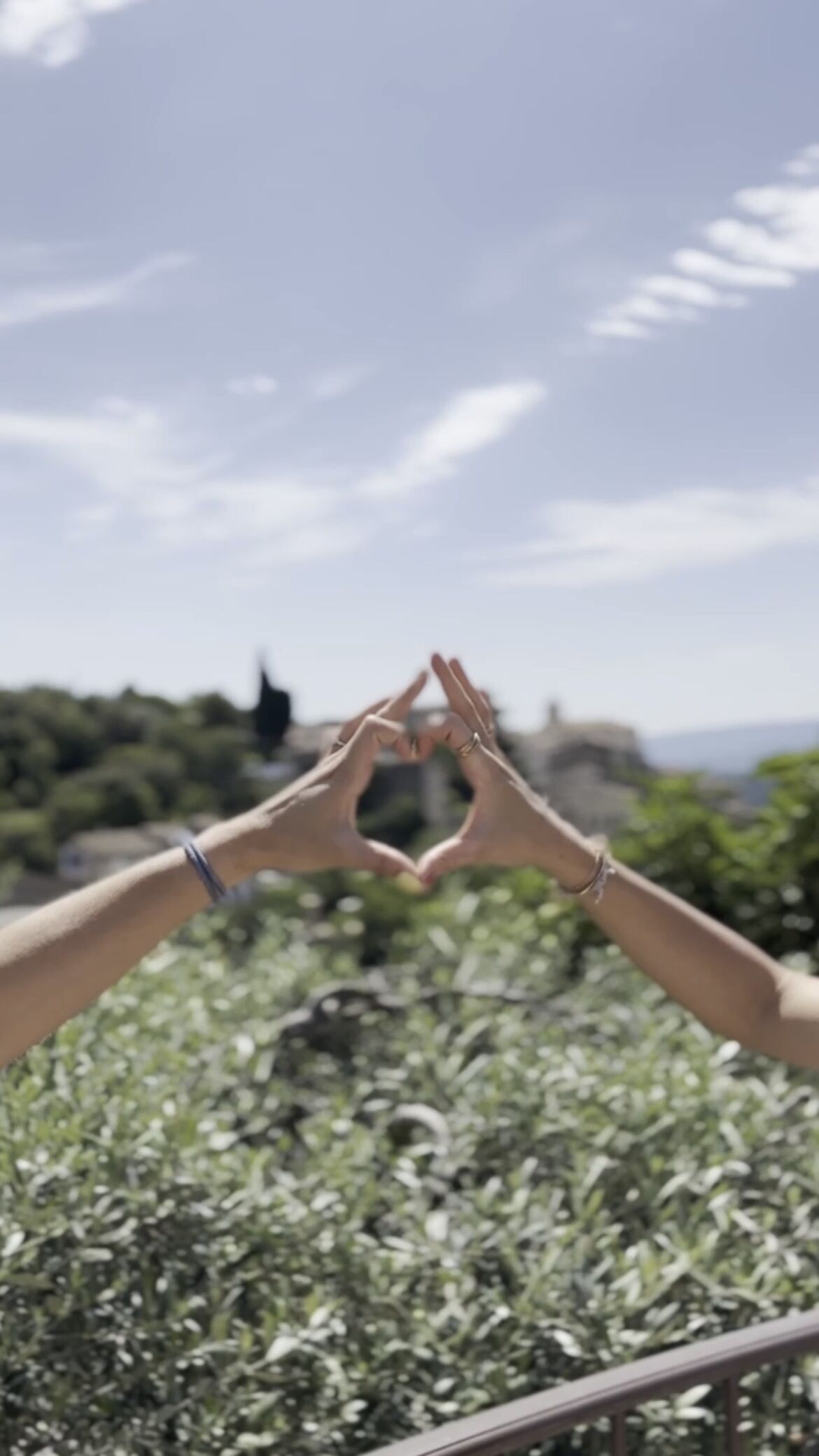 Two hands forming a heart shape against a backdrop of greenery and a clear sky.