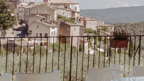 A terrace with wicker chairs and a view of a historic town with stone buildings and a mountain in the background.