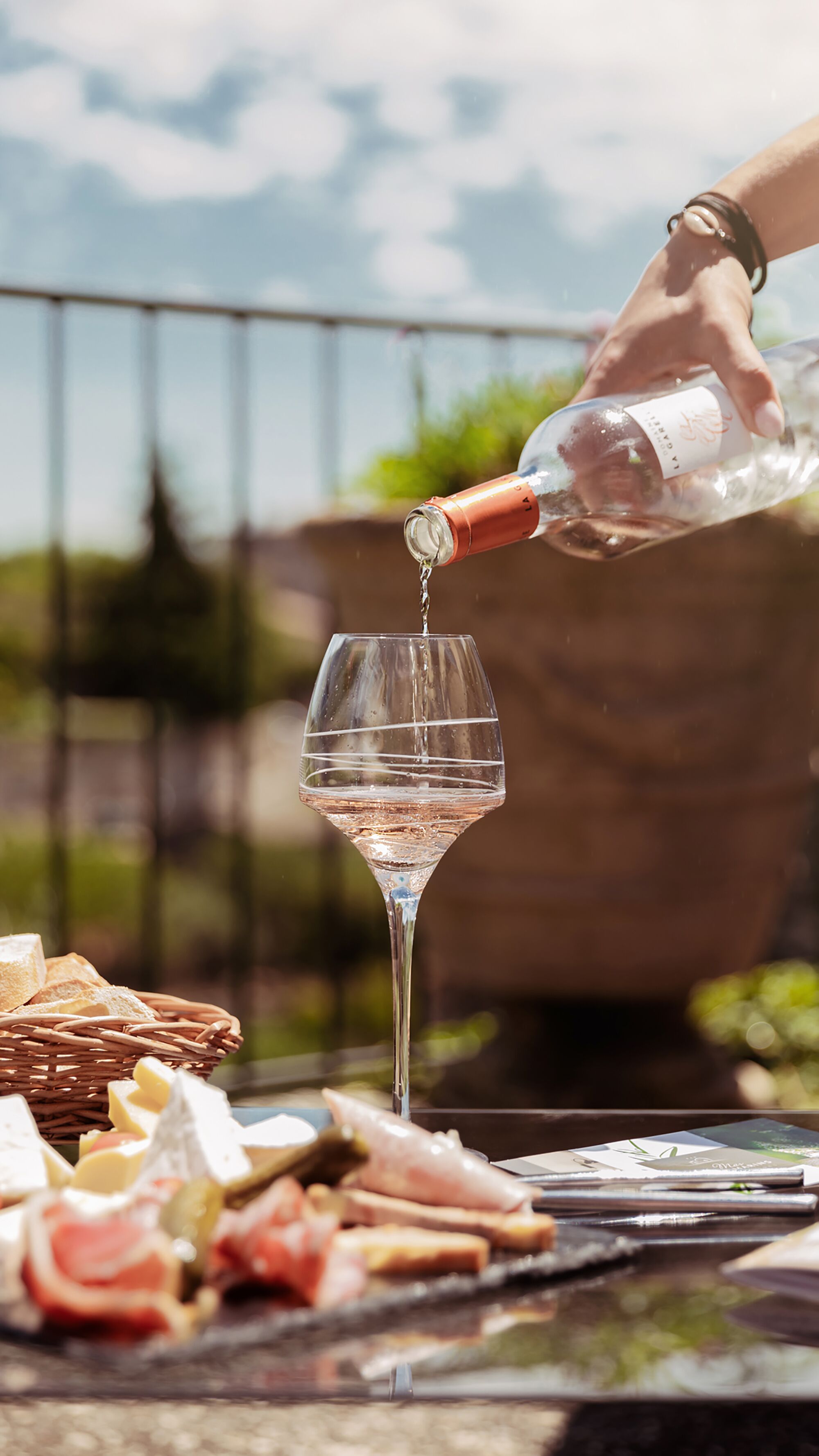 A hand pouring a clear liquid into a wine glass, with a wicker basket and food items in the foreground.