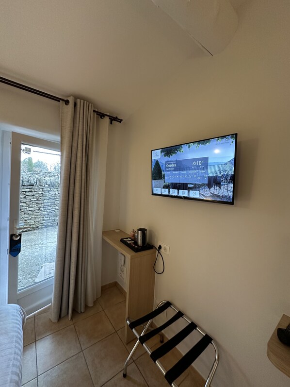 A room with a TV displaying a weather forecast, a window with curtains, a small wooden desk, and a metal chair. - Mas des Romarins, The Originals Relais (Gordes)