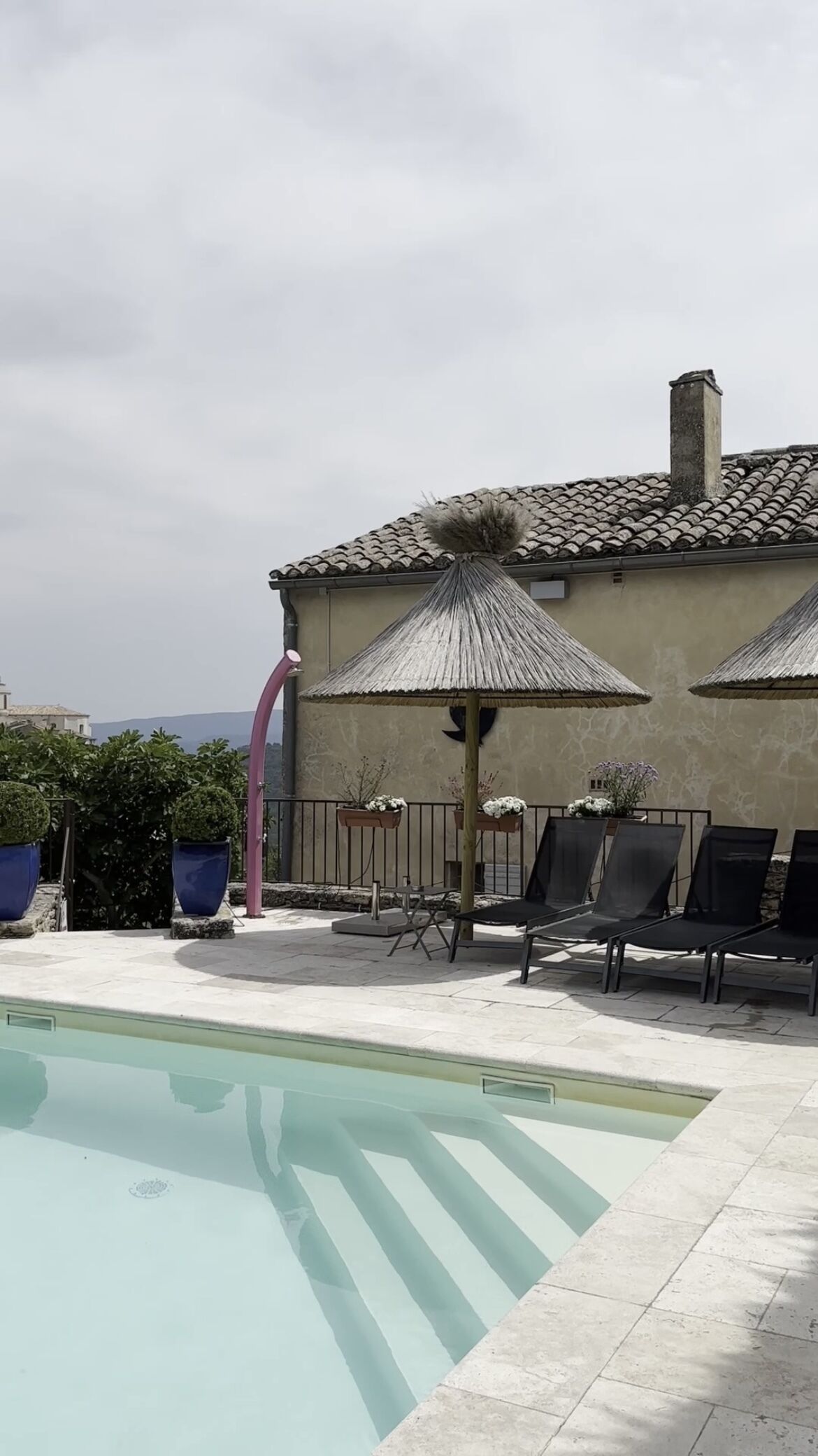 Una piscina con sillas de descanso y sombrillas, un muro de piedra y un edificio con techo de tejas.