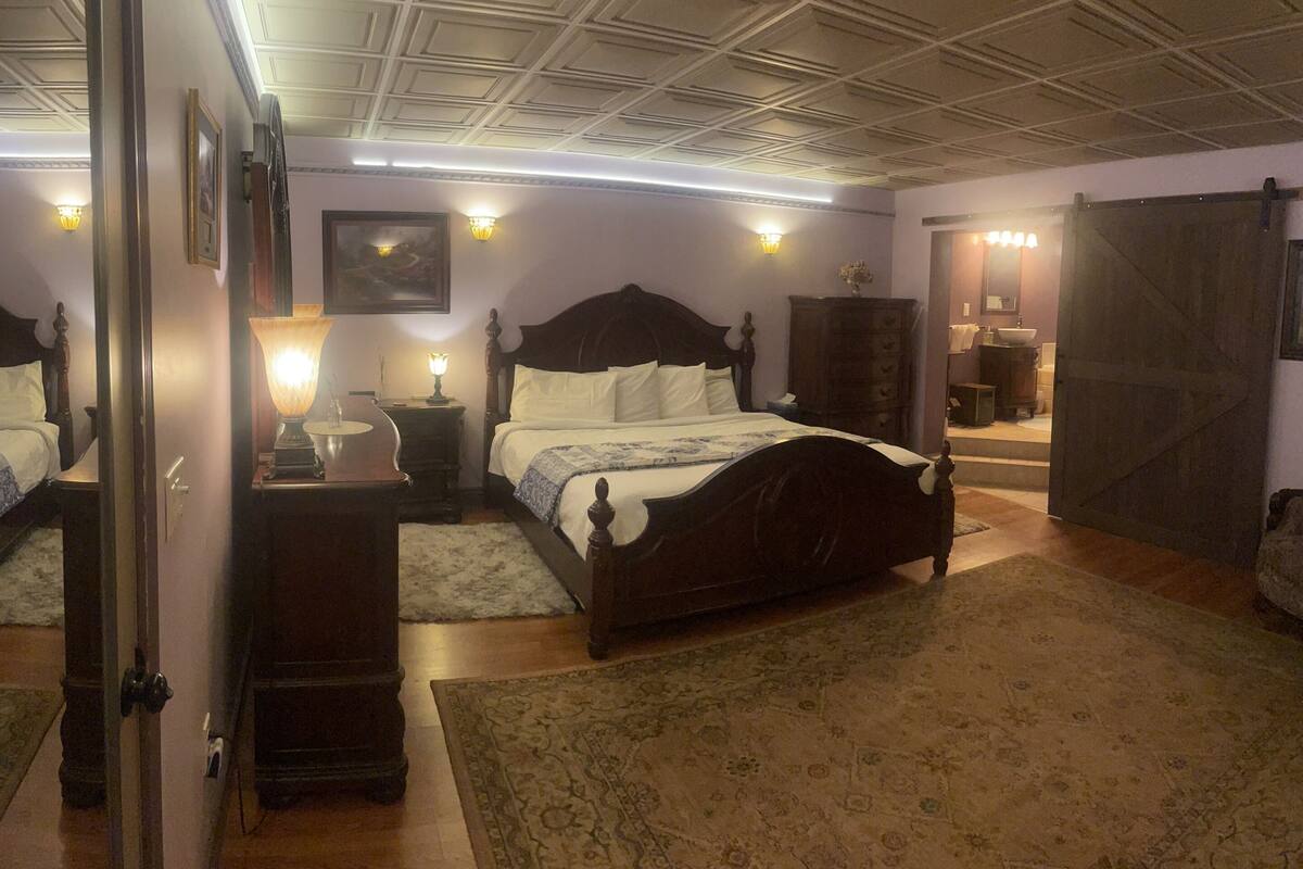 A bedroom with a large bed, a dresser, a nightstand, a mirror, and a door leading to a bathroom.