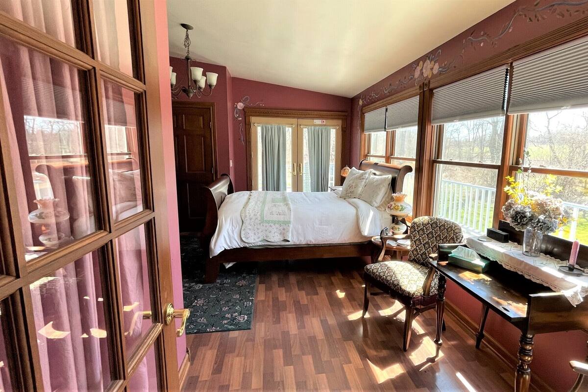 A bedroom with a bed, a desk, and a chair. There is a window with blinds, a door, and a chandelier.