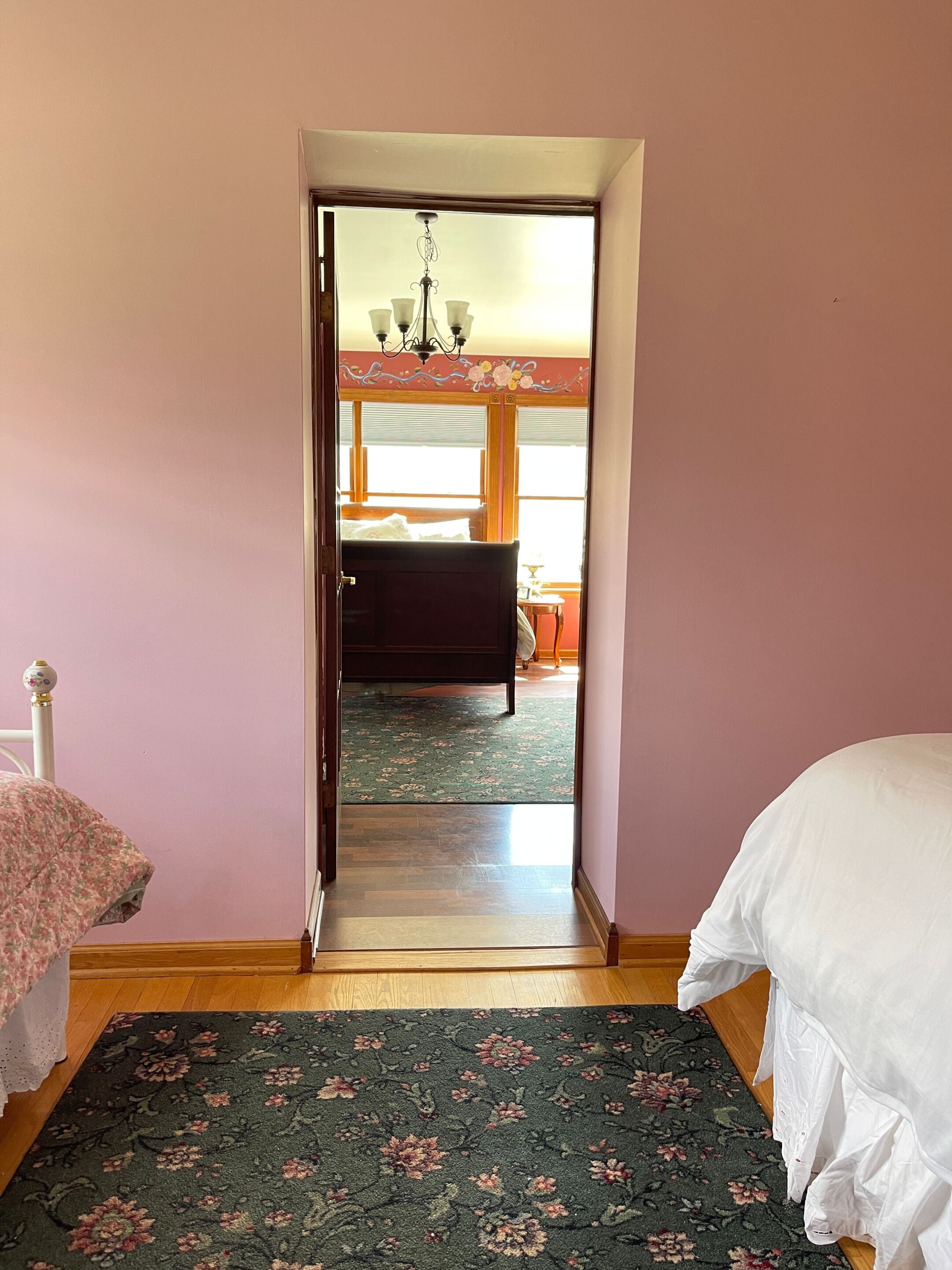 A bedroom with a bed, a floral-patterned rug, and a view into another room with a window and a chandelier.