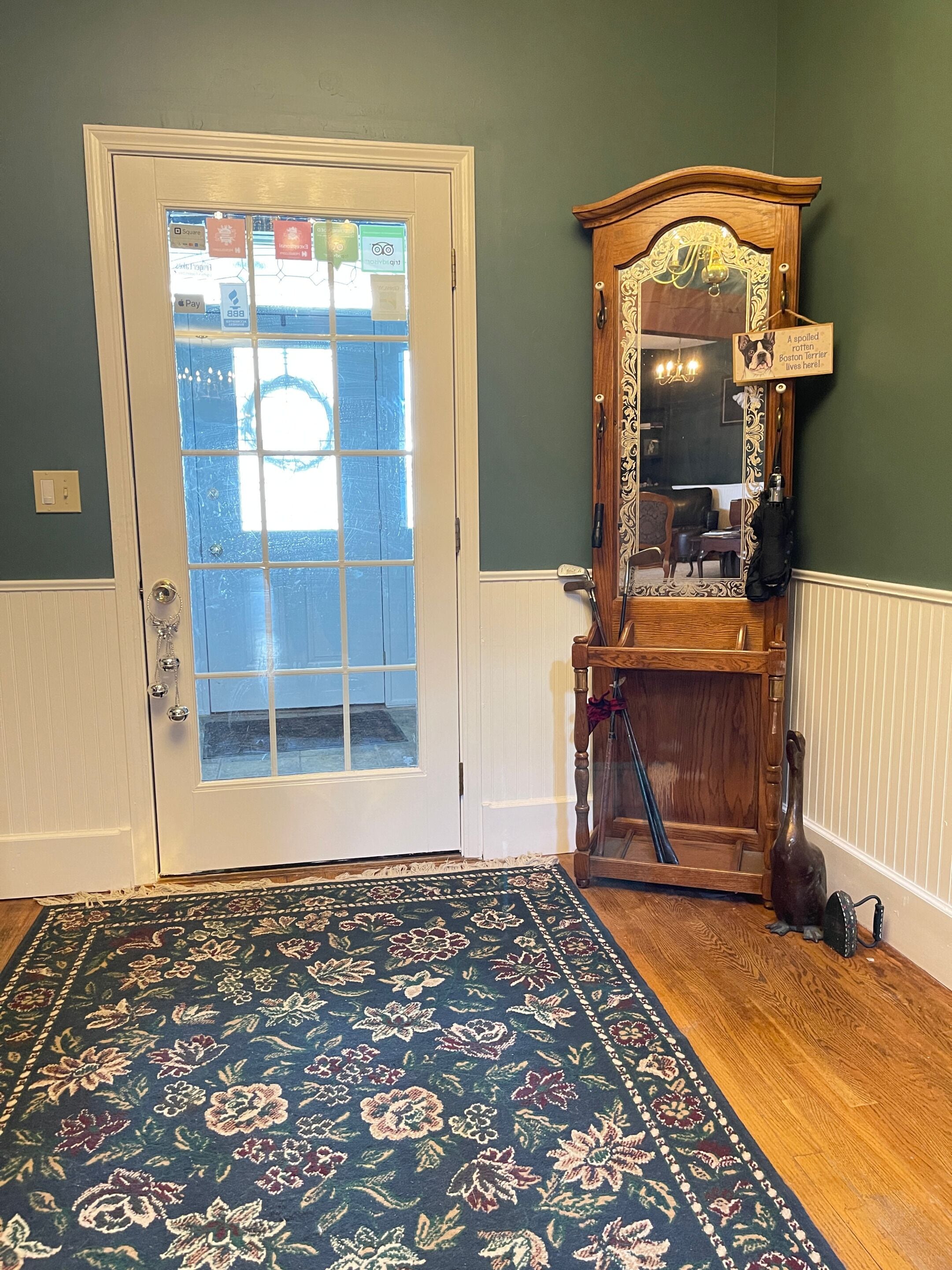 A room with a wooden floor, a floral patterned rug, a white door with frosted glass panels, and a wooden mirror with a dog sign.