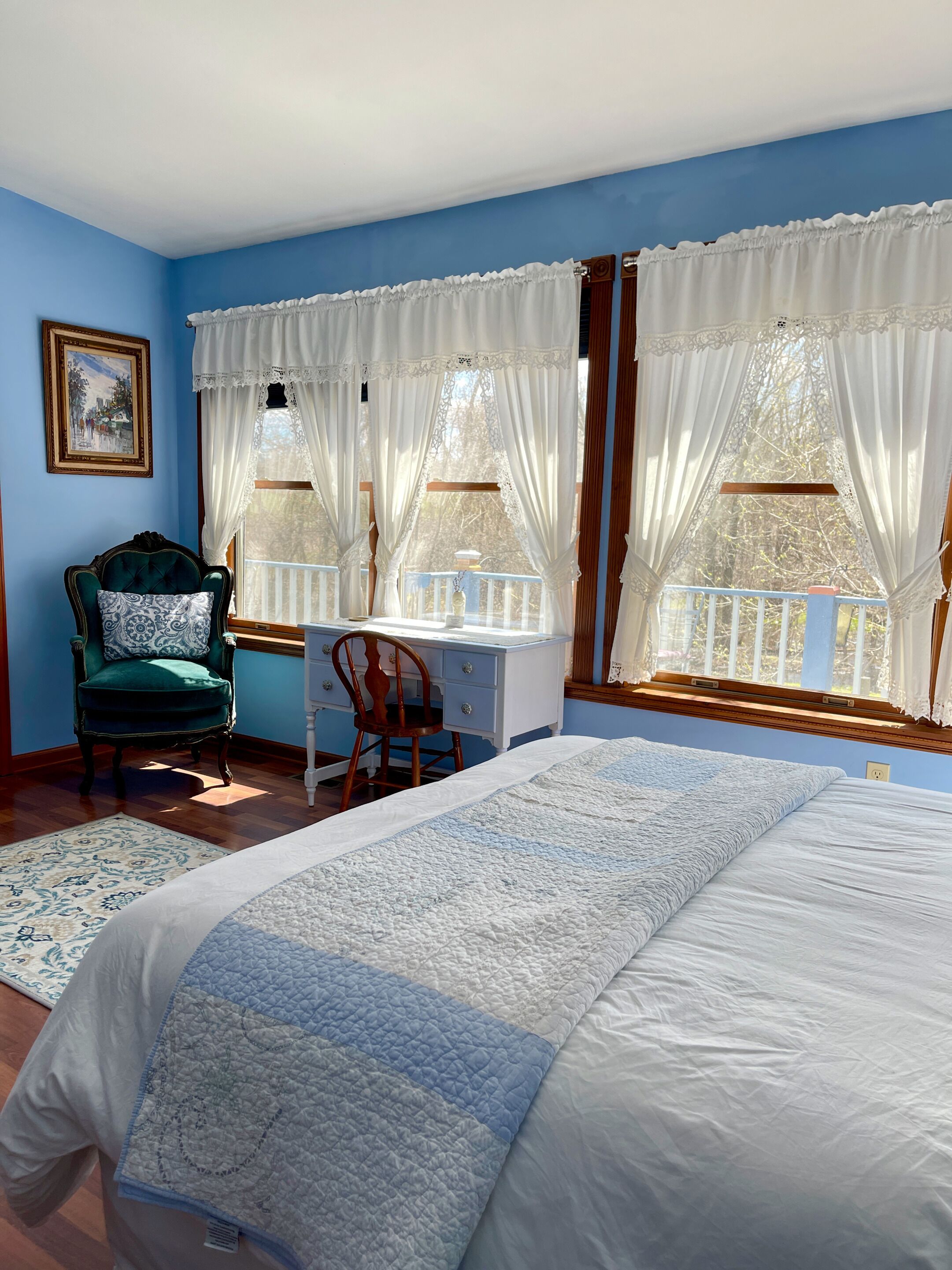 A bedroom with a large bed, a desk, and a chair. There are two windows with white curtains.