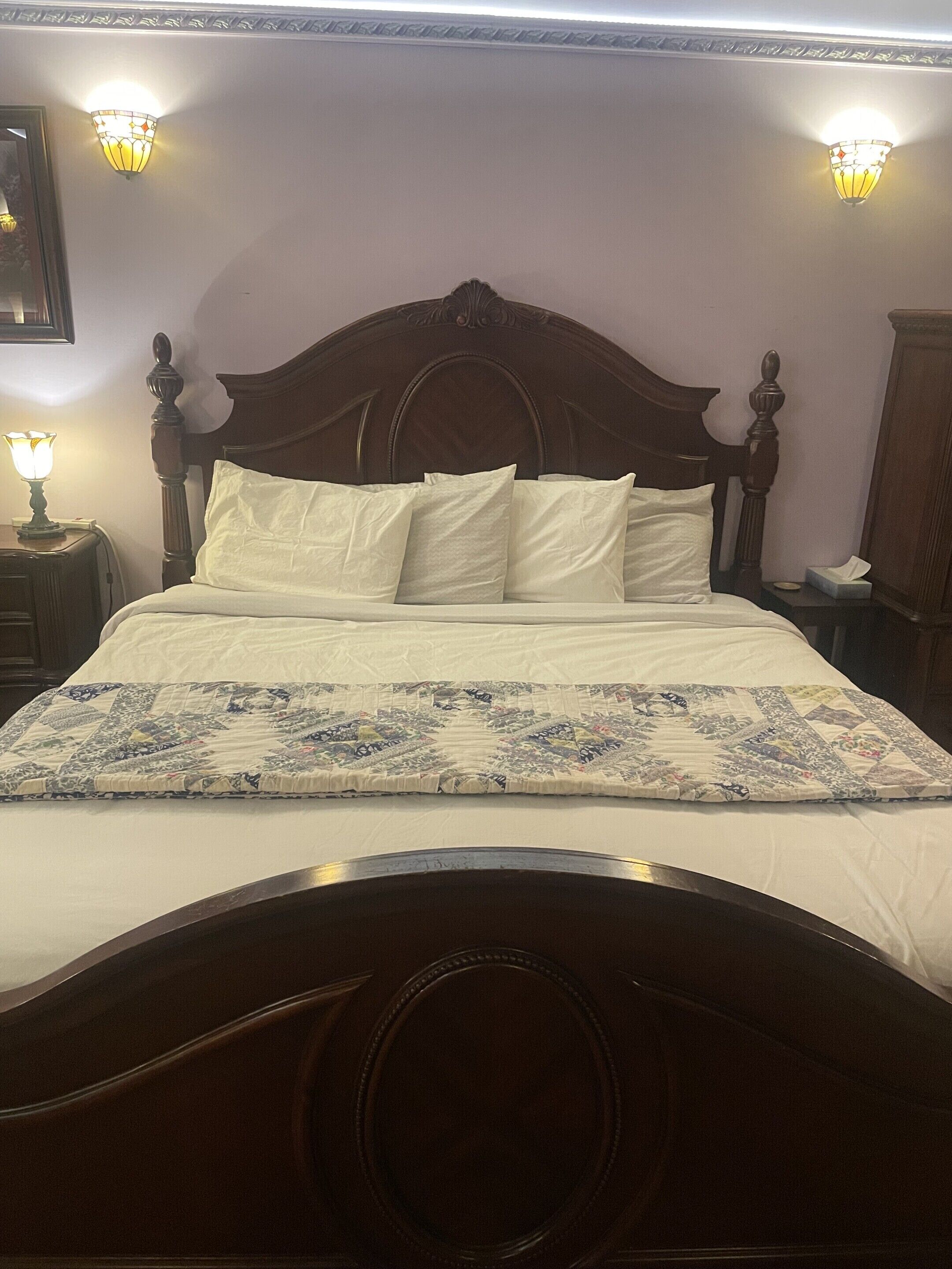 A bed with a wooden headboard, white bedding, and a quilt. There are two wall sconces on either side of the bed.