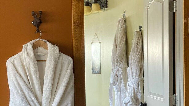 A bathroom with a wooden door, a towel rack with a white towel, and a mirror with a wall-mounted light. - The Yorkshire Inn (Phelps)