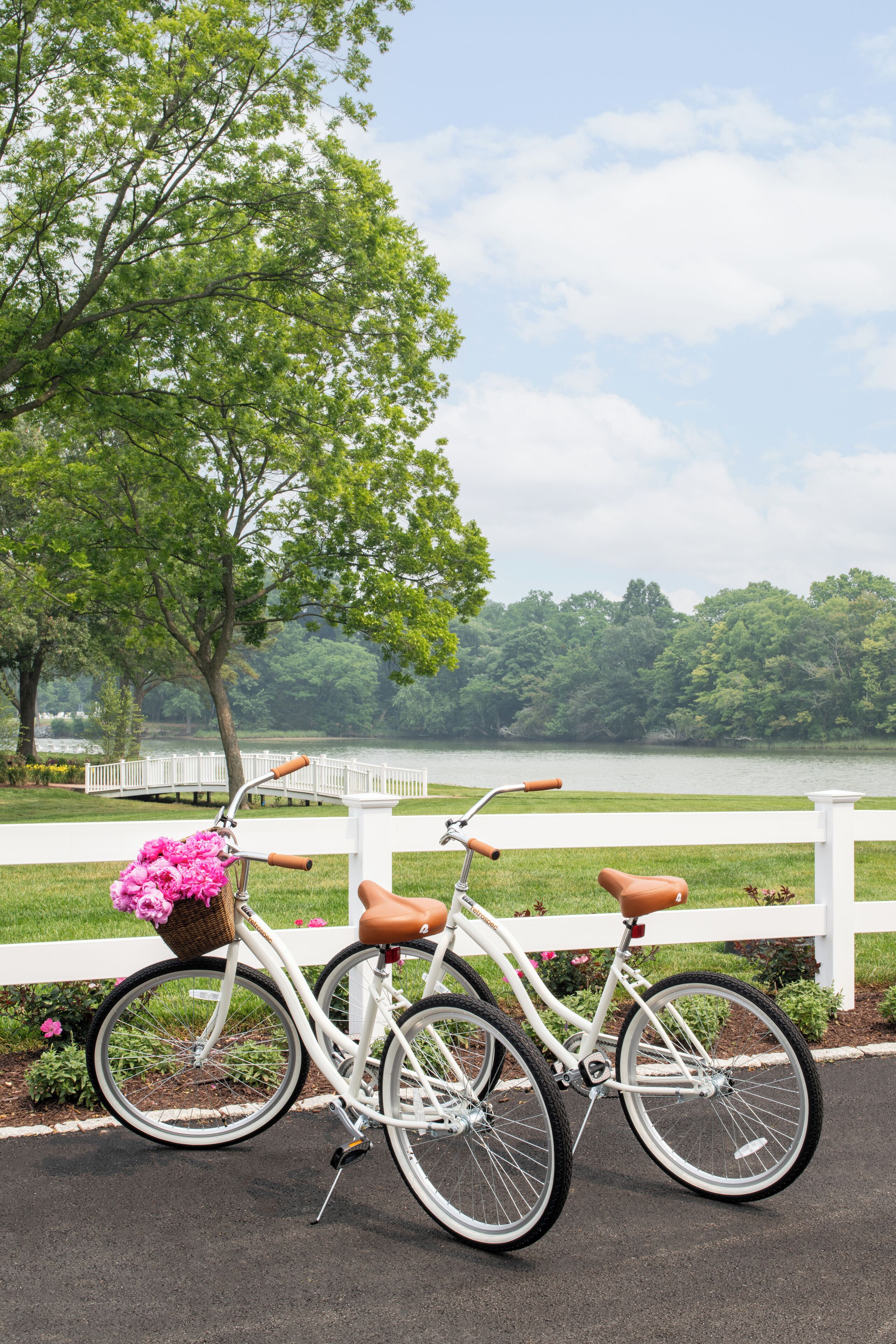 Una bicicleta blanca con una cesta de flores rosas estacionada junto a una valla blanca cerca de un lago.