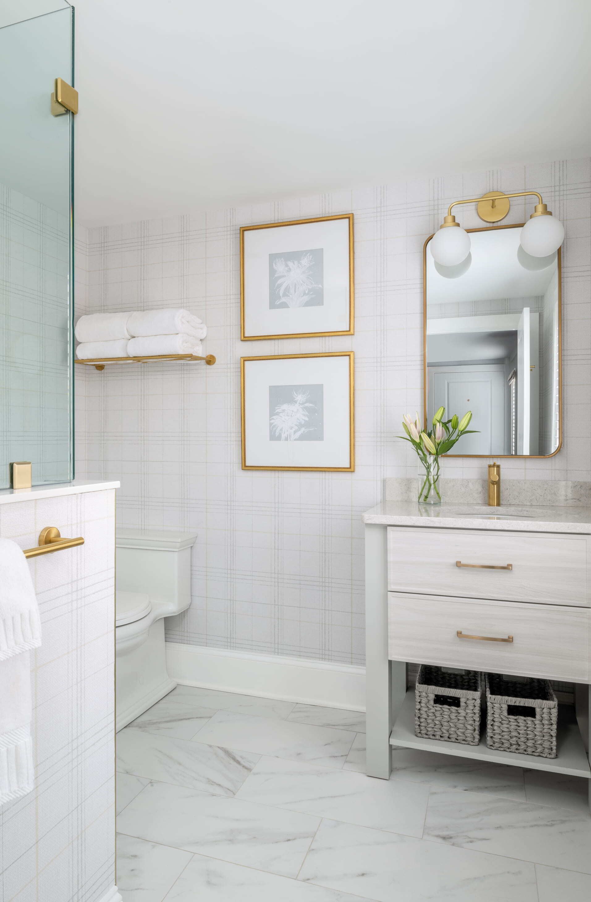 A bathroom with a white marble floor, a white vanity with gold handles, a gold-framed mirror with lights, and two framed pictures on the wall.