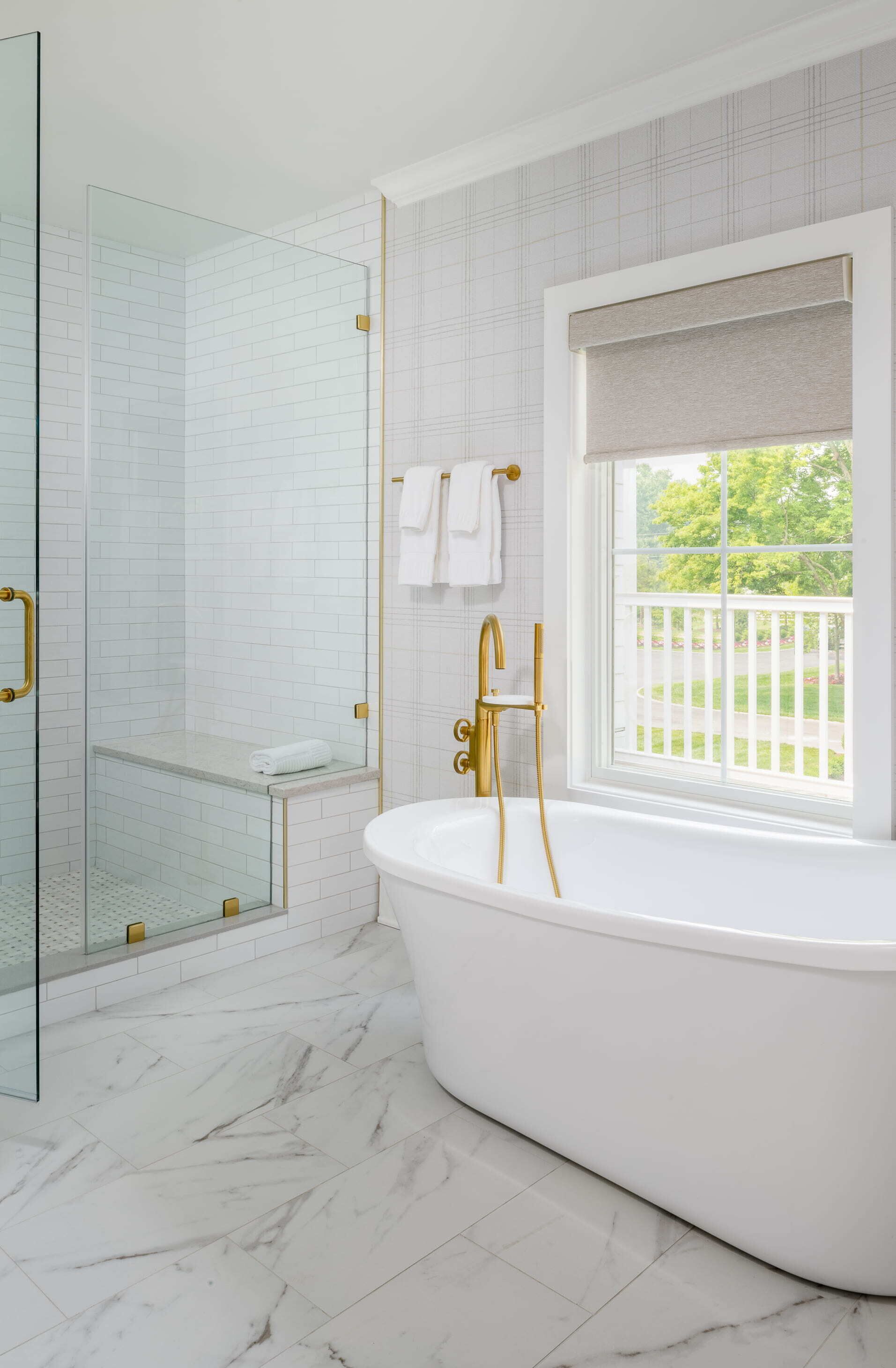 A bathroom with a freestanding bathtub, a glass shower enclosure, and a window with a view of a white fence and greenery.