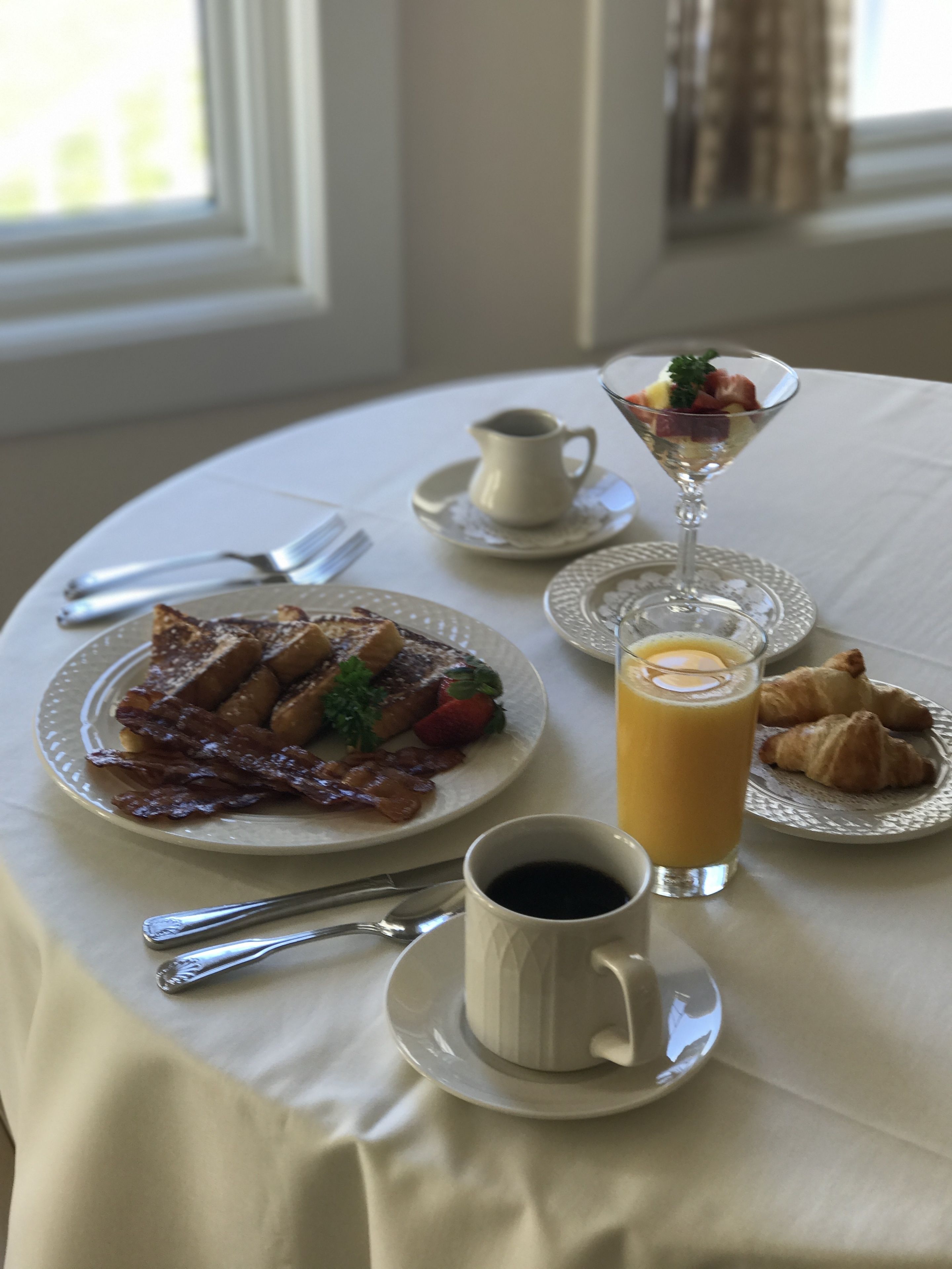 A breakfast table with croissants, a glass of orange juice, a cup of coffee, and a plate of eggs with bacon and strawberries.