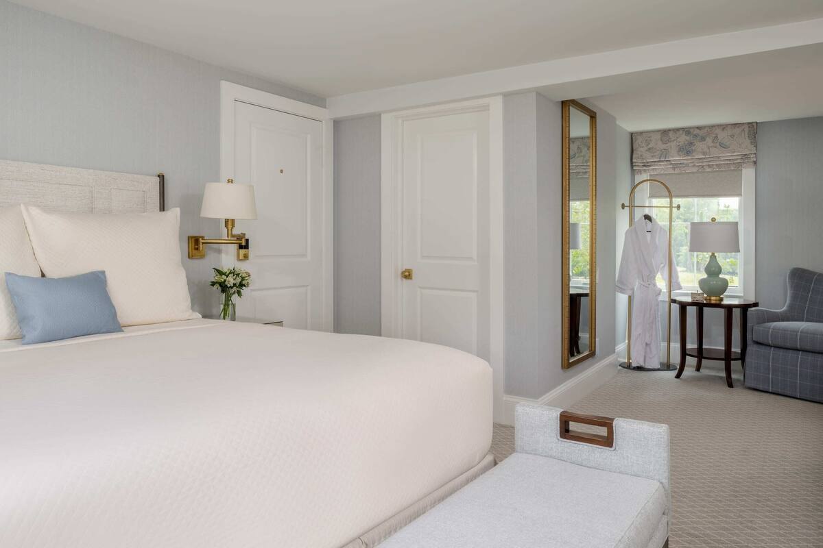 Ein Hotelzimmer mit einem großen Bett, einer Sitzecke mit einem Sessel, einem kleinen Tisch und einem Spiegel.