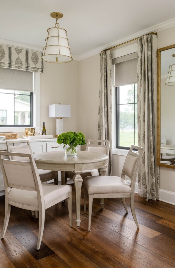 A dining room with a round table, white chairs, a vase of green flowers, a hanging light fixture, and a window with curtains.