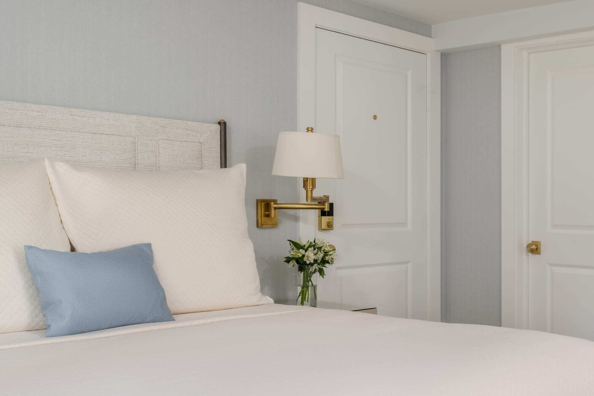 A neatly made bed with white and blue pillows, a gold bedside lamp, and a vase with flowers on a nightstand.