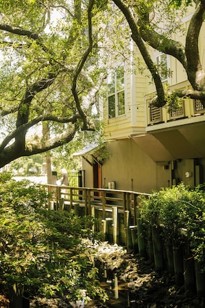 A wooden bridge over a stream with lush greenery and a building in the background.