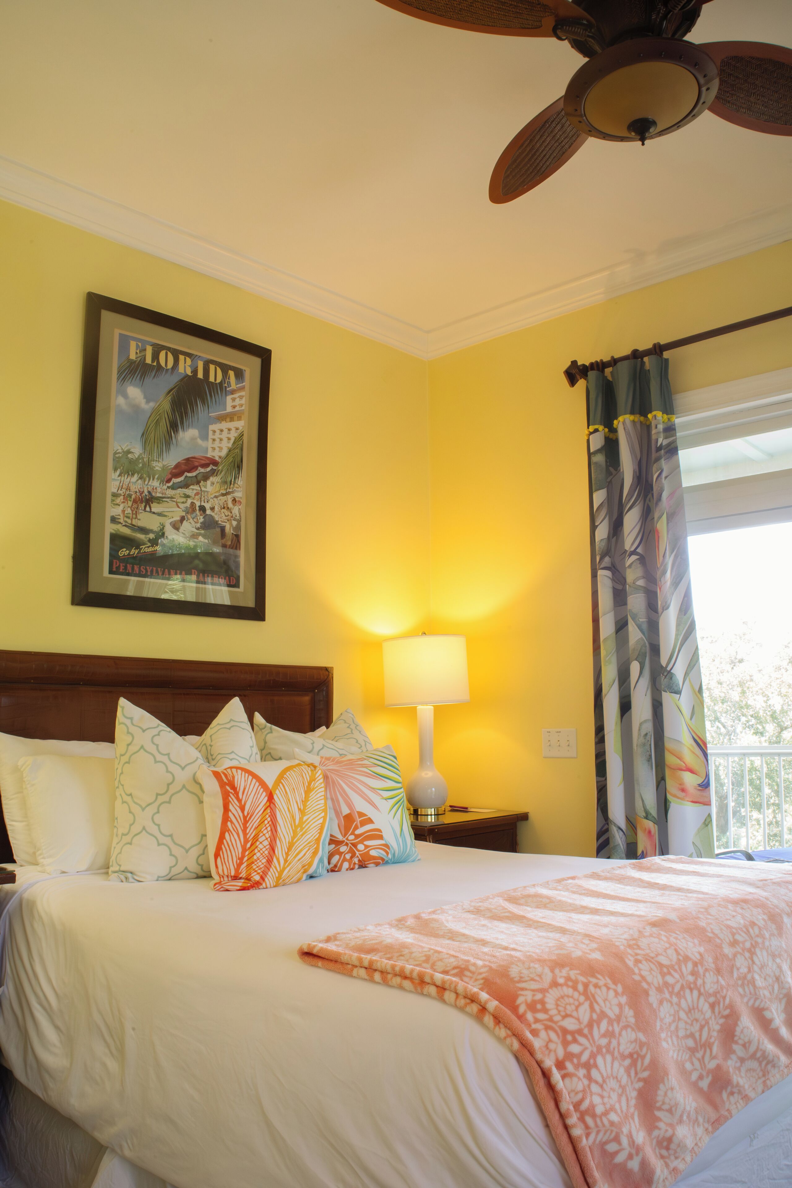 Une chambre d’hôtel avec un lit, un ventilateur de plafond, une lampe, une fenêtre avec des rideaux et un tableau encadré au mur.