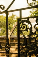 Una reja de hierro forjado con una estructura de madera detrás, un letrero con texto y vistas a árboles.