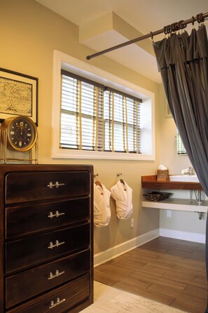 A bathroom with a wooden dresser, a framed map, a window with blinds, and a shower curtain. - Water's Edge Inn - Adults Only (Folly Beach)