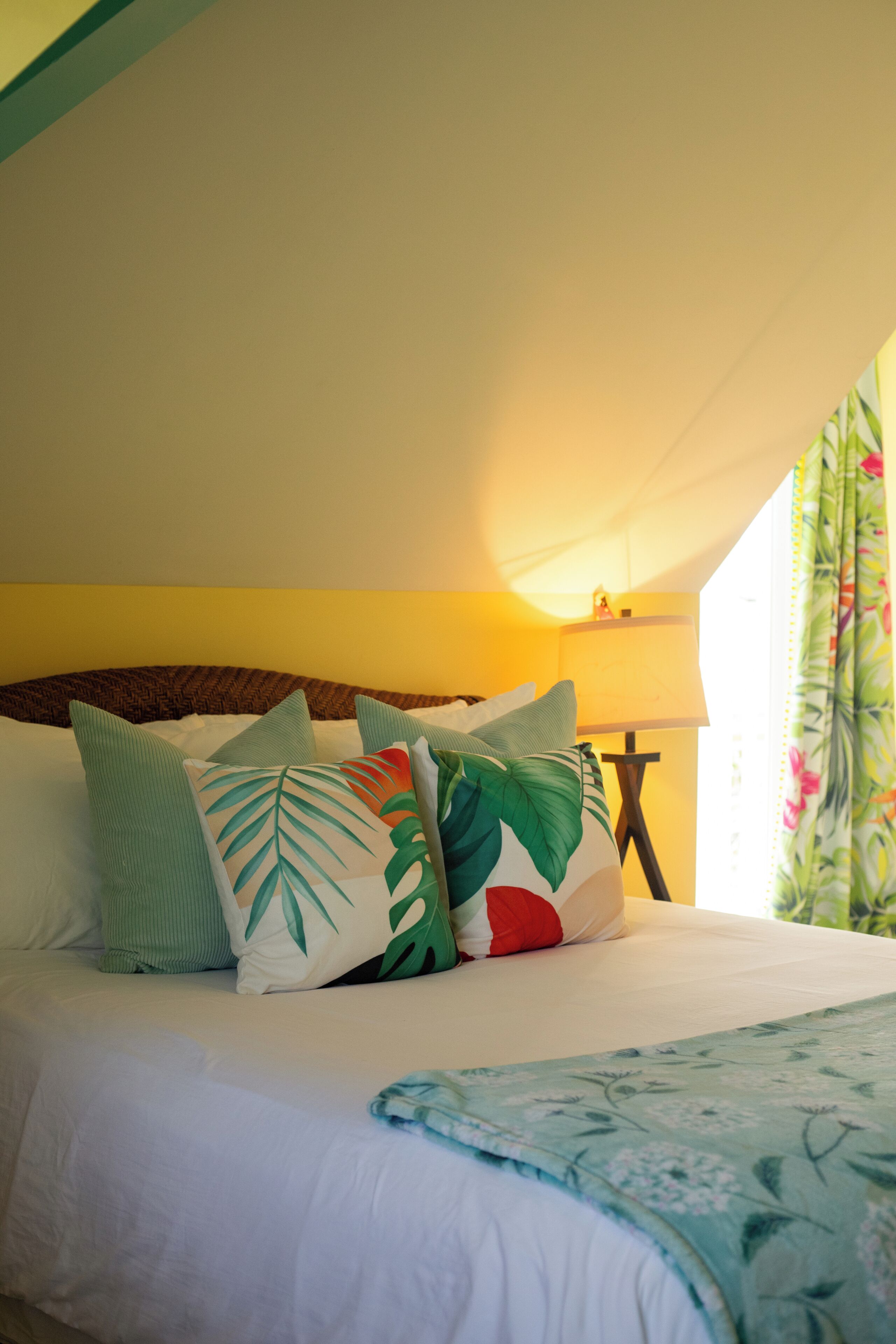 A bed with multiple pillows, a bedside table with a lamp, and a window with curtains.