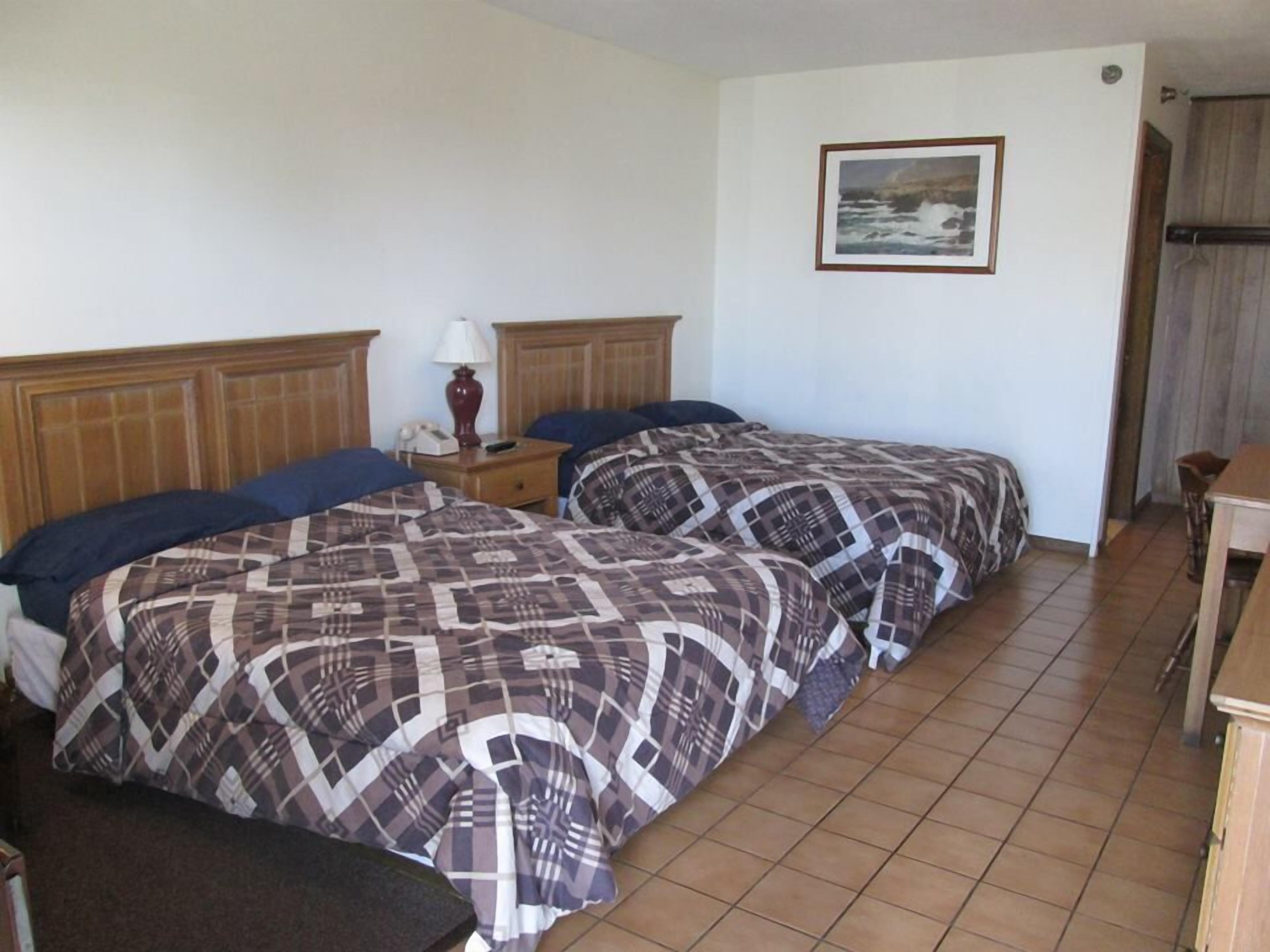 Quarto de hotel com duas camas, cabeceira de madeira, mesa de cabeceira com abajur, quadro emoldurado na parede e mesa de madeira.