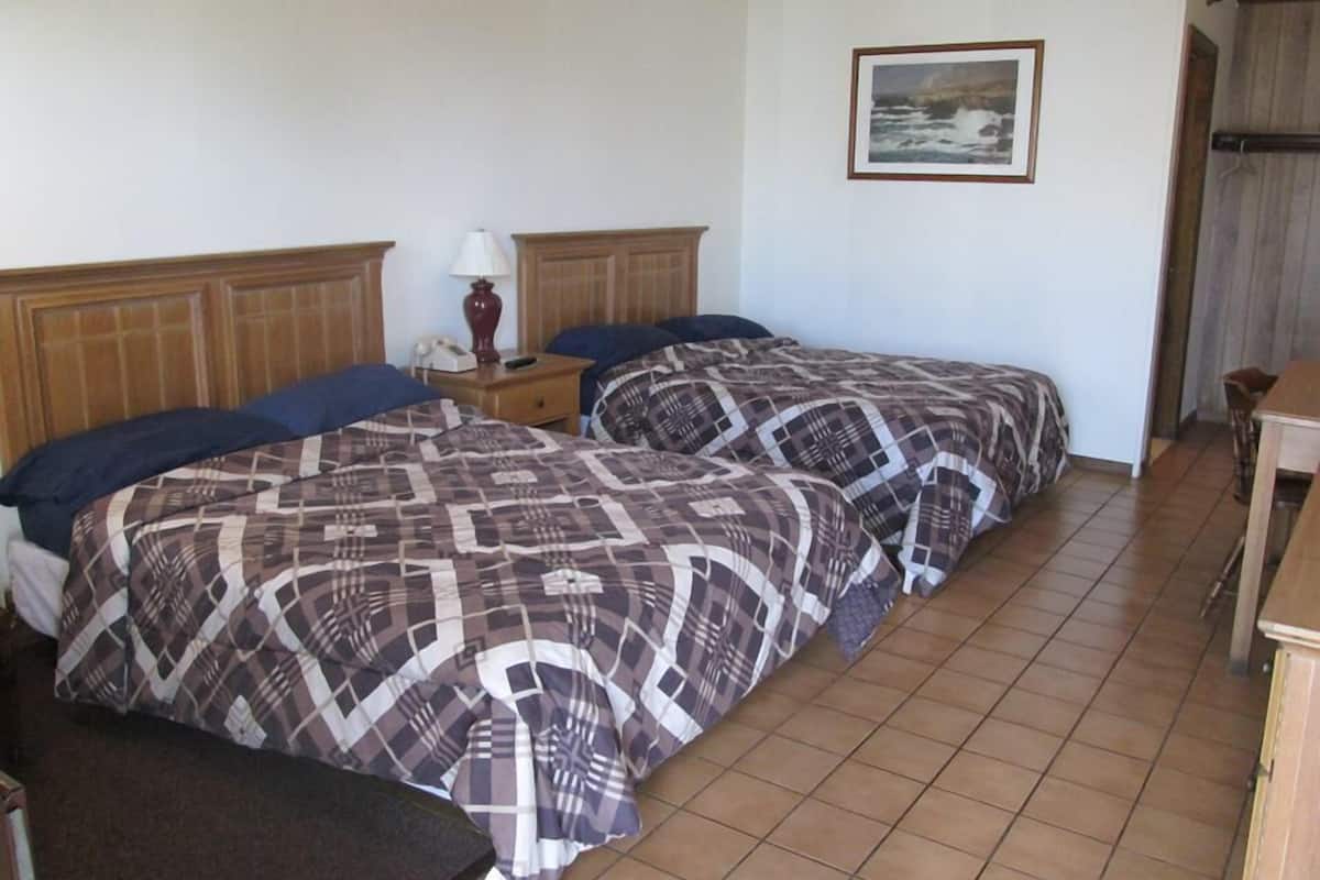 A hotel room with two beds, a wooden headboard, a nightstand with a lamp, a framed picture on the wall, and a wooden table.