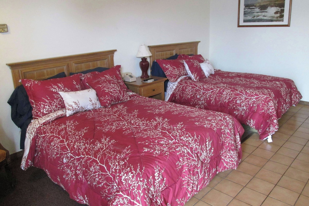 A room with two beds, each with red bedding and white tree patterns, a wooden headboard, a nightstand with a lamp, and a framed picture on the wall.