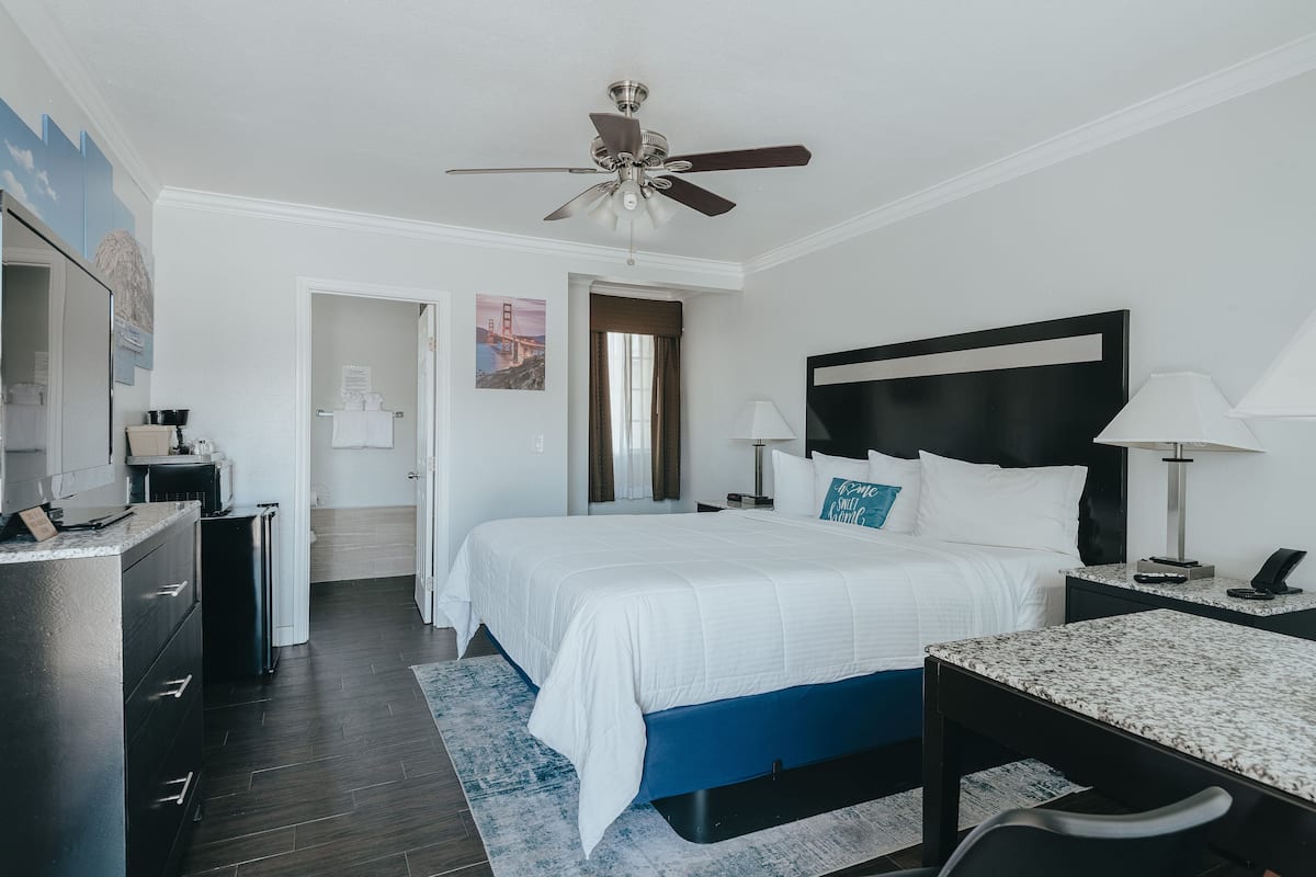 Habitación de hotel con cama, escritorio, silla, ventilador de techo, mesita de noche, lámpara, cómoda y puerta al baño.