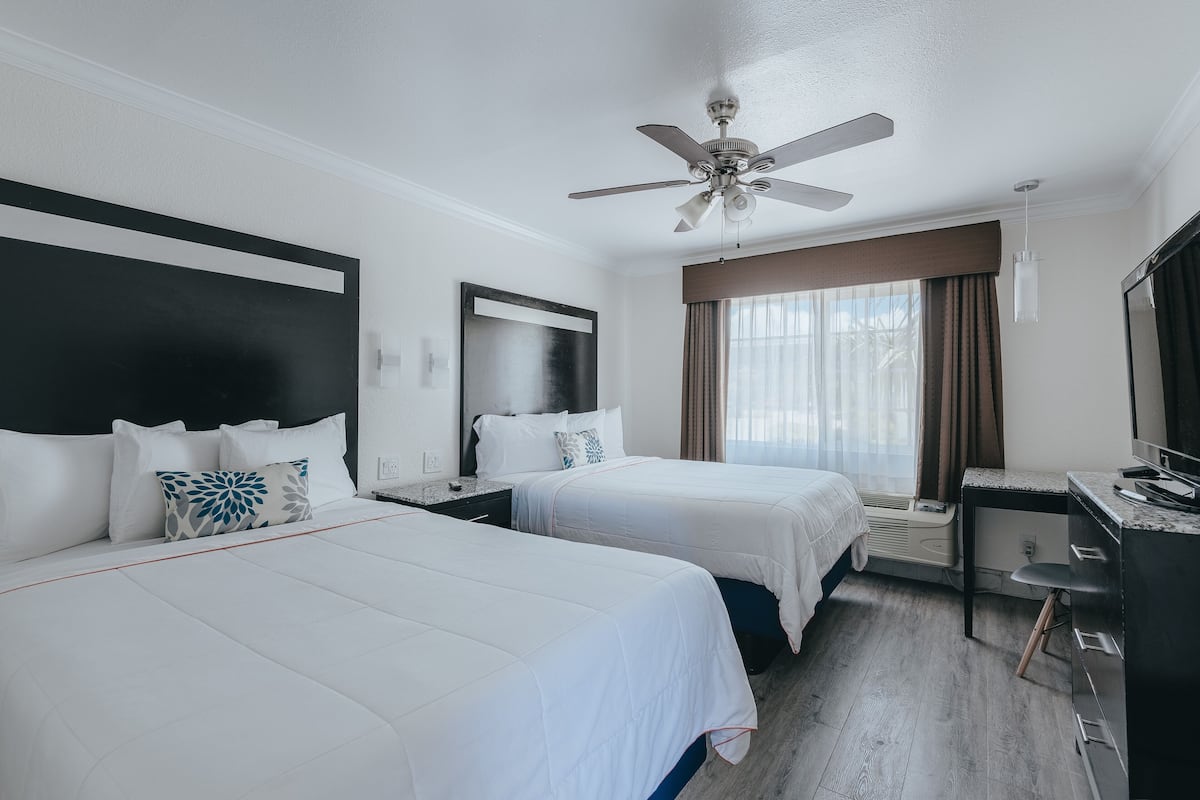 Habitación de hotel con dos camas, un escritorio, una silla, un ventilador de techo y un ventanal con cortinas.