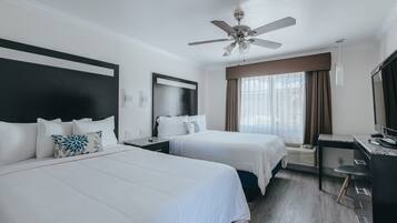 Quarto de hotel com duas camas, uma escrivaninha, uma cadeira, ventilador de teto e uma janela grande com cortinas.