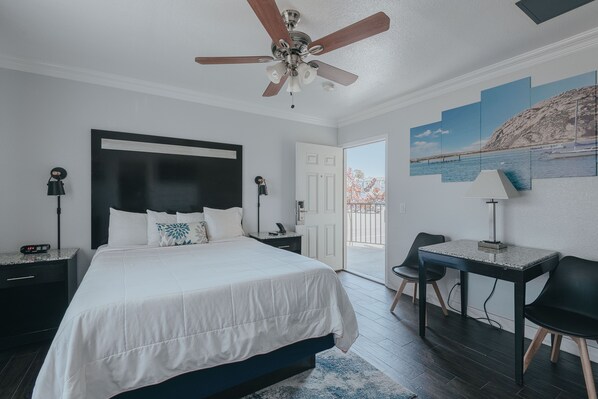 A bedroom with a bed, a desk, a chair, a ceiling fan, and a view of the outdoors.