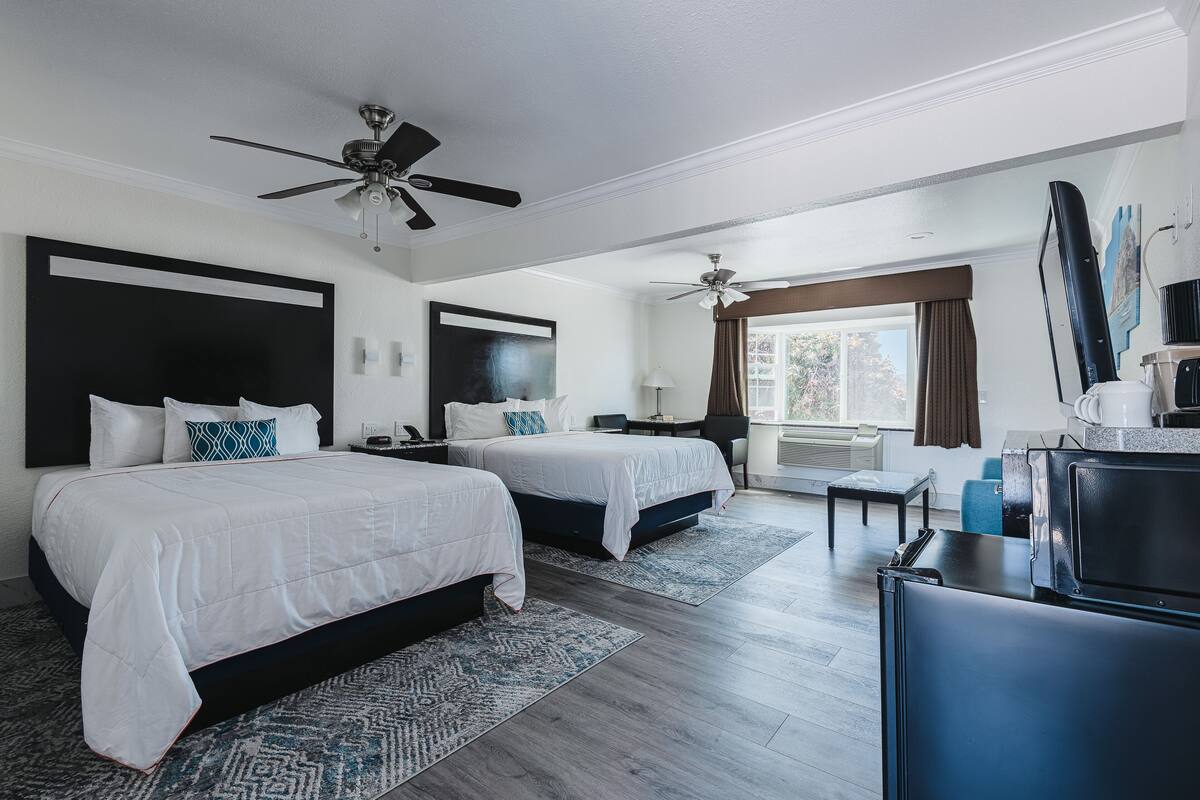 Habitación de hotel con dos camas, un escritorio, ventilador de techo, ventana grande con cortinas y televisor de pantalla plana.