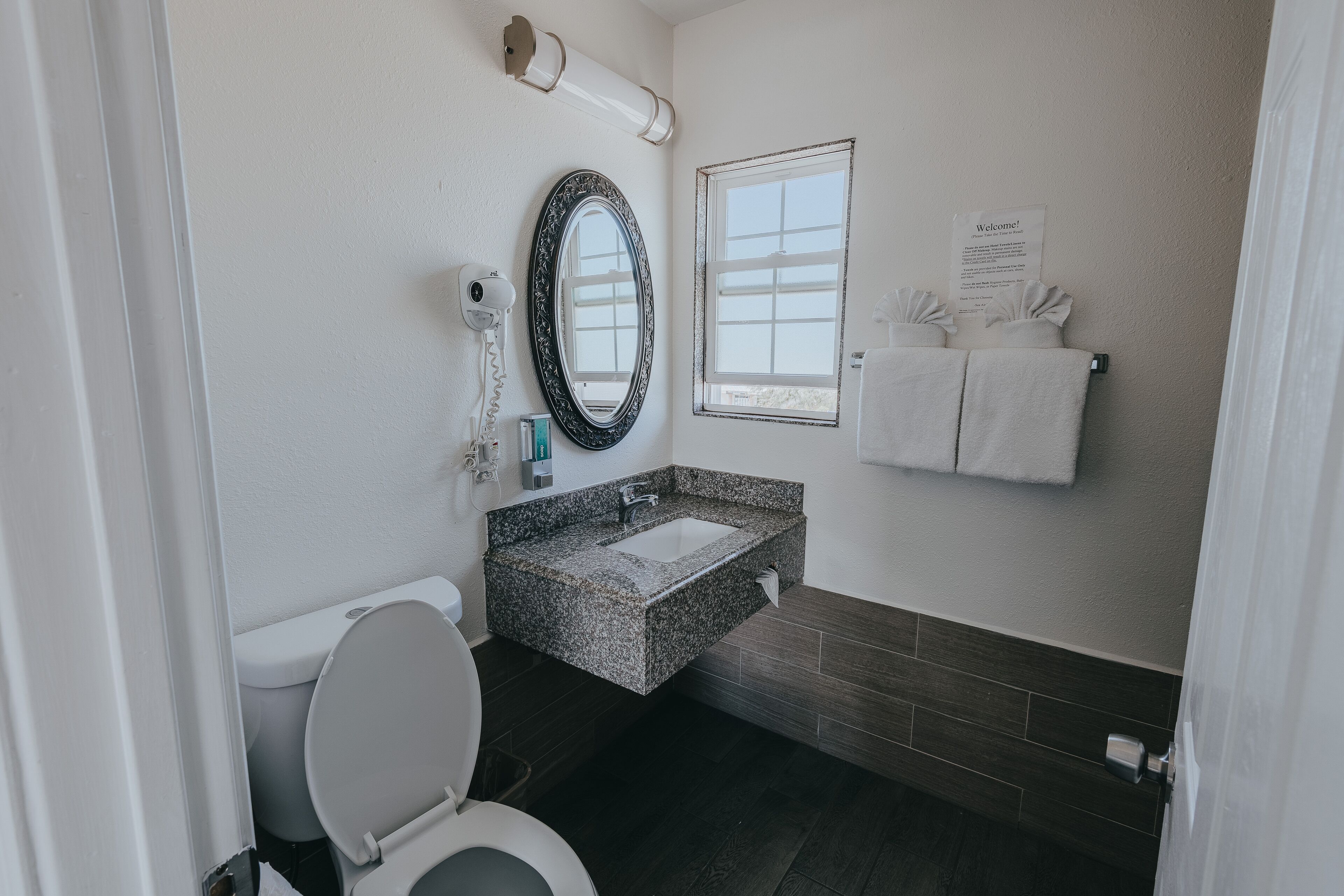 Un baño moderno con inodoro, lavamanos con encimera de granito, espejo grande, ventana y toallero con toallas.