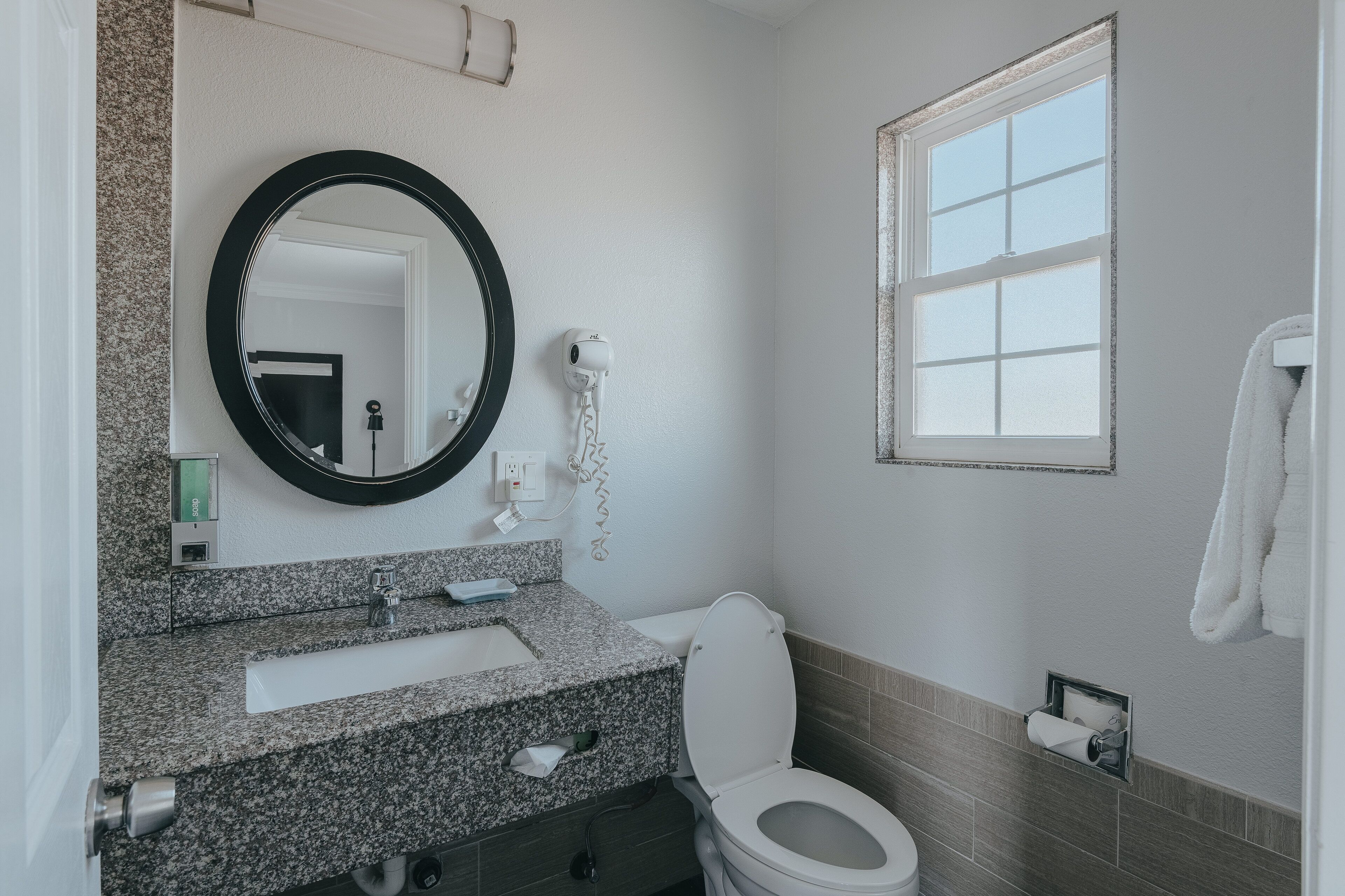 A bathroom with a granite countertop, a white toilet, a round mirror, a hairdryer, and a window with blinds.