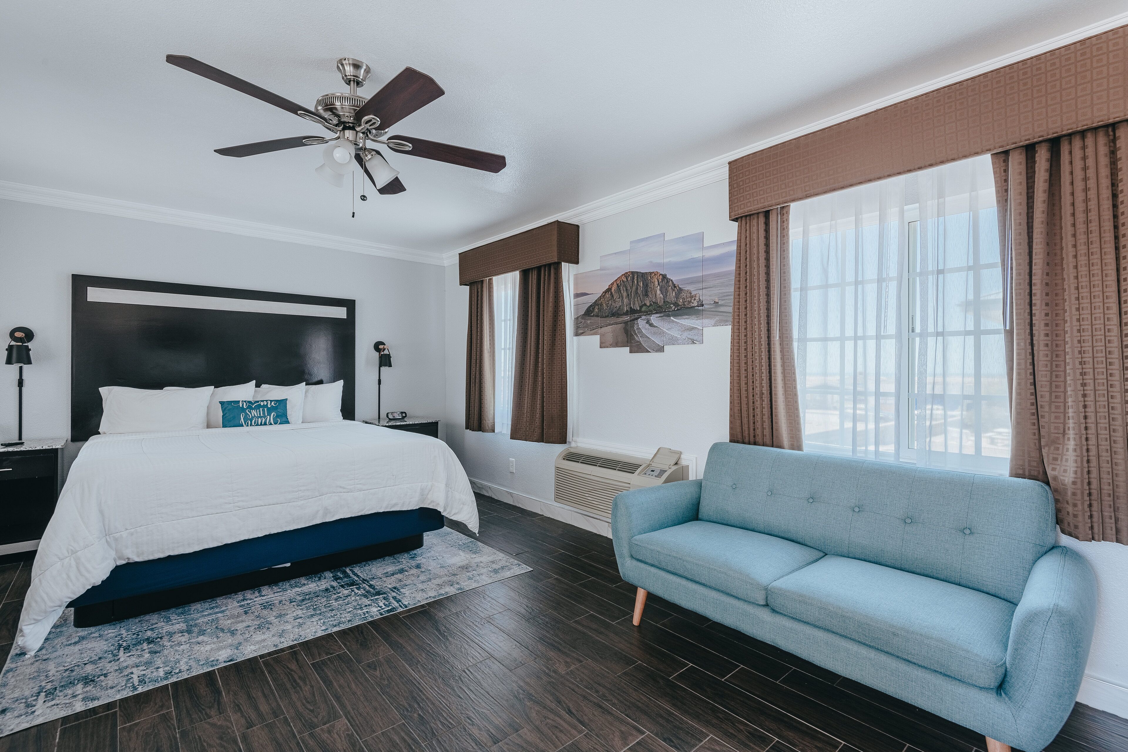 A bedroom with a bed, a blue sofa, a ceiling fan, a large window with curtains, and a wall-mounted TV.