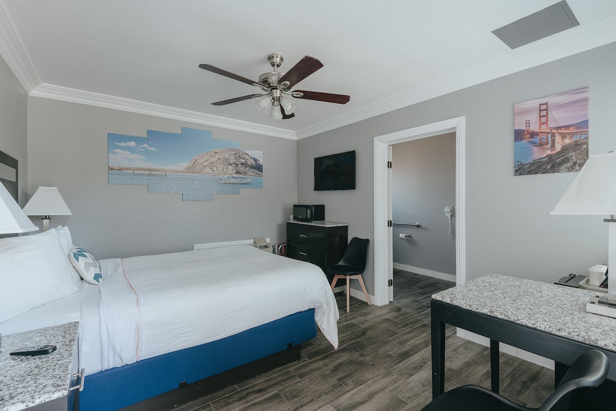 Habitación de hotel con cama, escritorio, silla, ventilador de techo, obras de arte y una puerta que da a otra habitación.