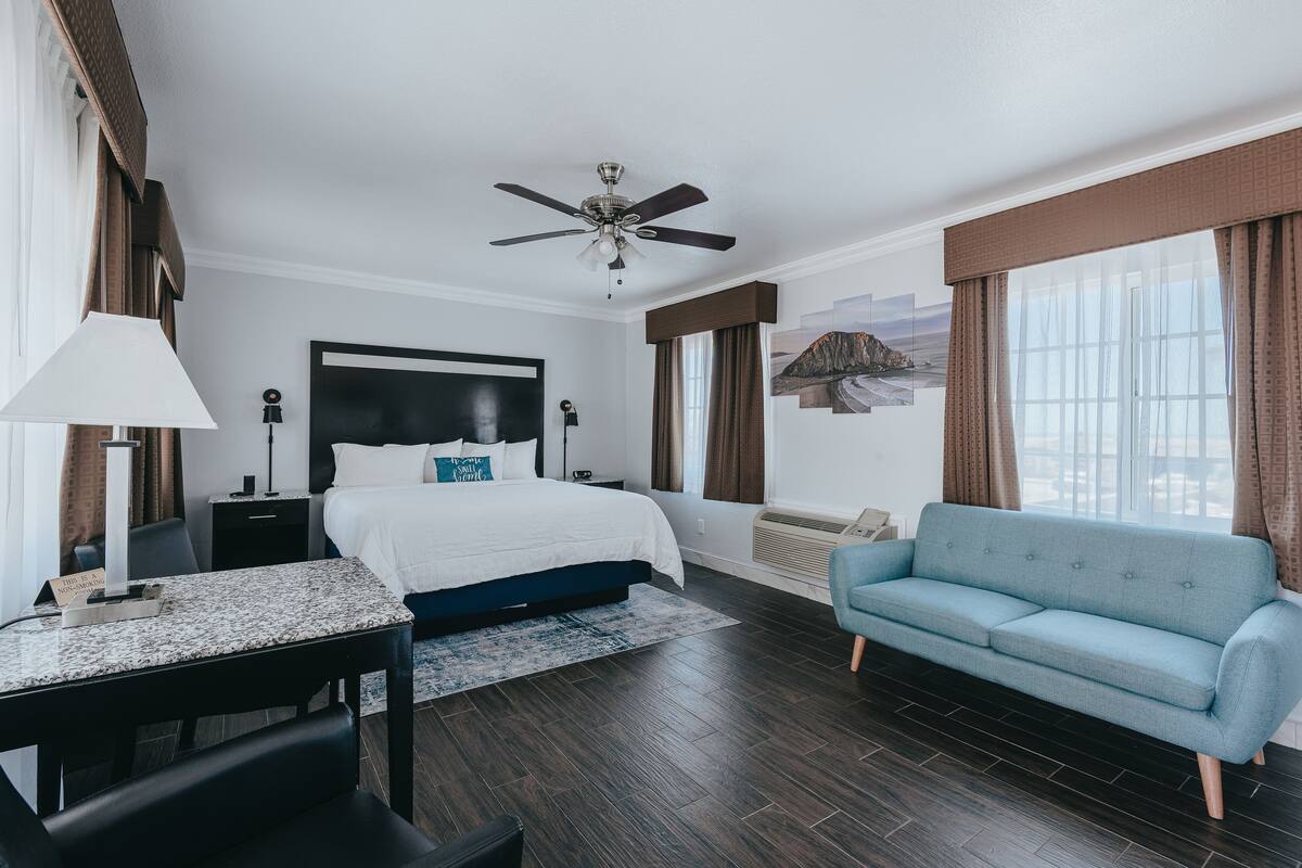 Habitación de hotel con cama, escritorio, sofá, ventilador de techo y una ventana grande con cortinas.