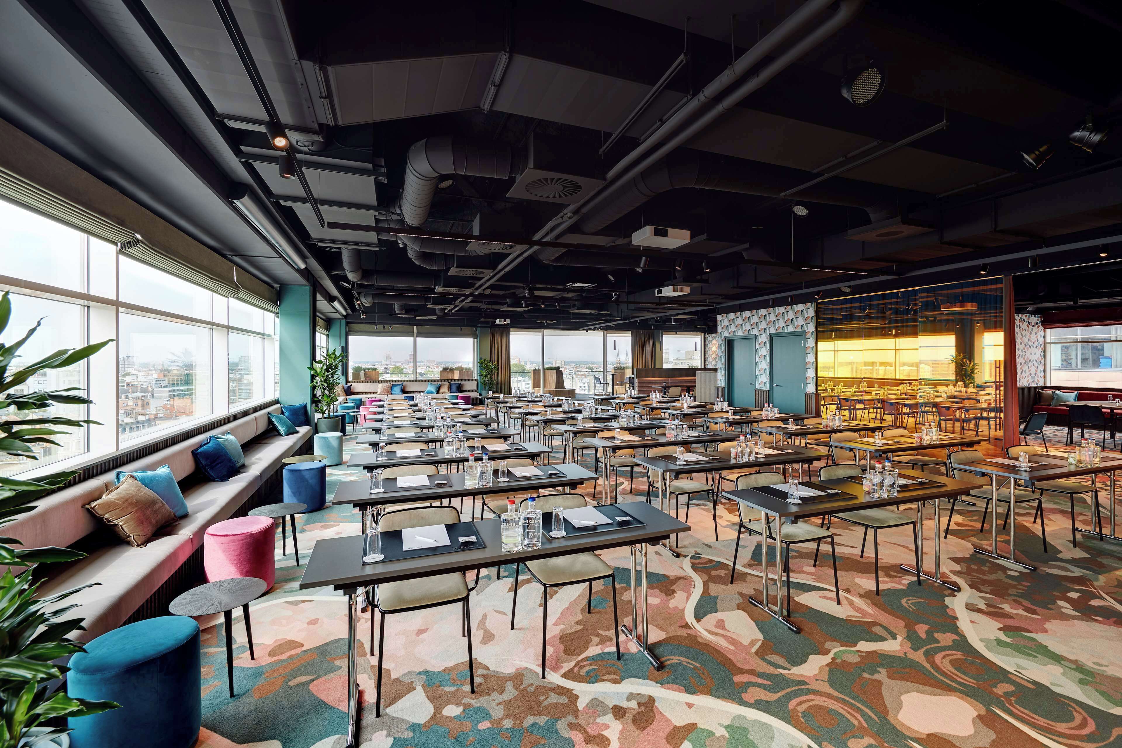 A spacious conference room with multiple tables and chairs, large windows offering a city view, and a seating area with cushions.