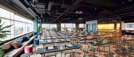 A spacious conference room with multiple tables and chairs, large windows offering a city view, and a seating area with cushions.
