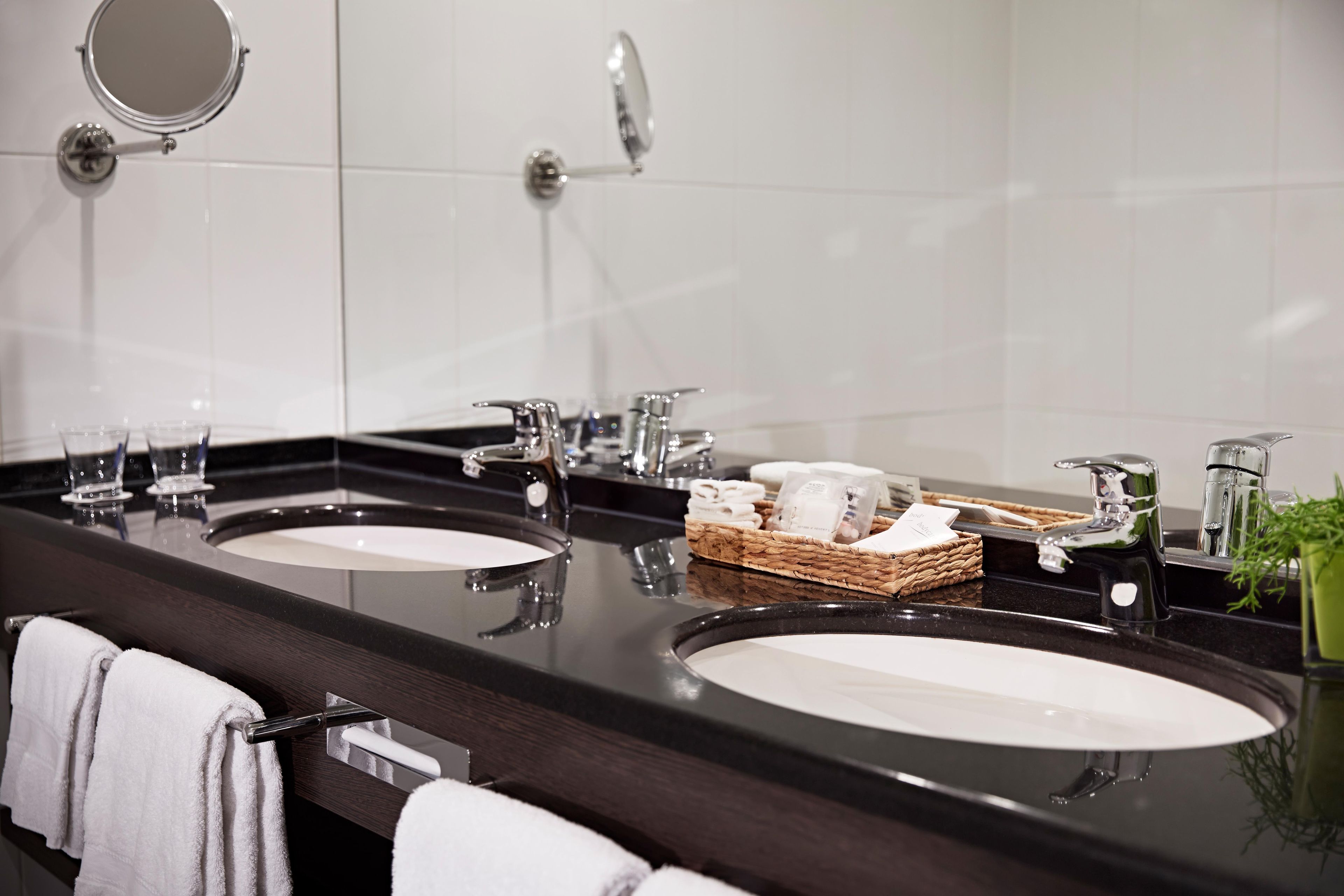 A modern bathroom with two sinks, a mirror, and a basket of toiletries.